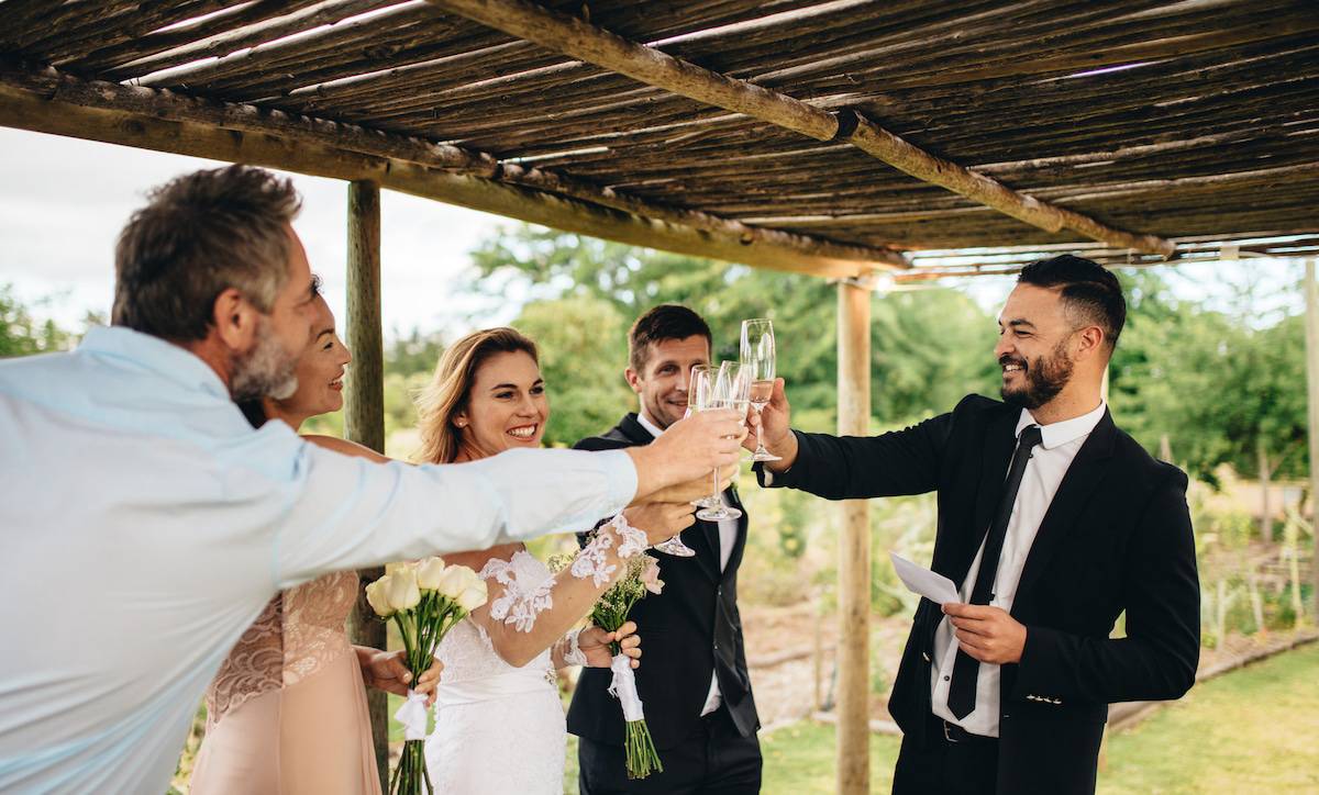 Guest toasting champagne with wedding couple. Young newlyweds clinking glasses and enjoying the moment with guest at wedding reception outside.