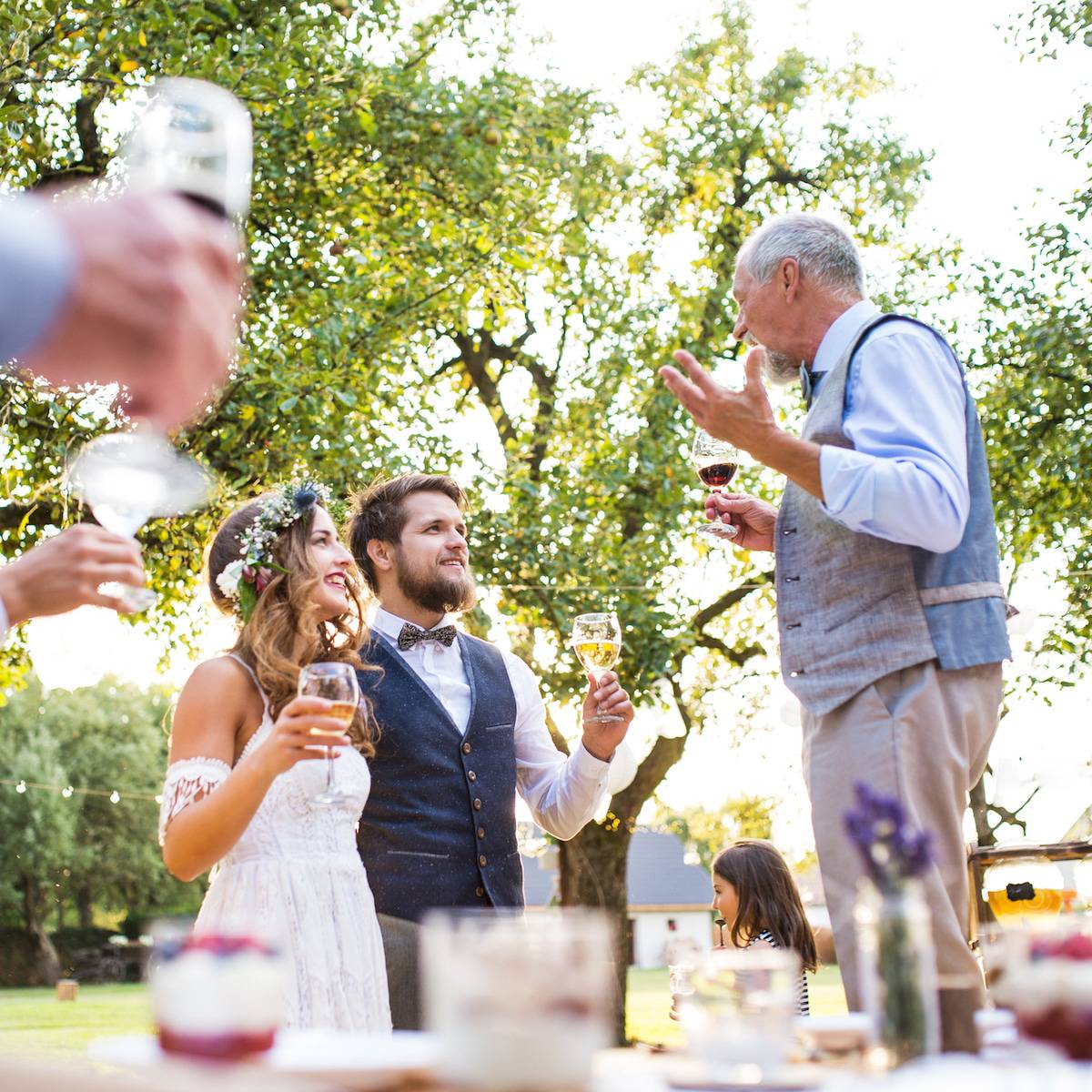 A senior man making speech at wedding reception outside in the background. Bride and groom and other guests holding glasses.