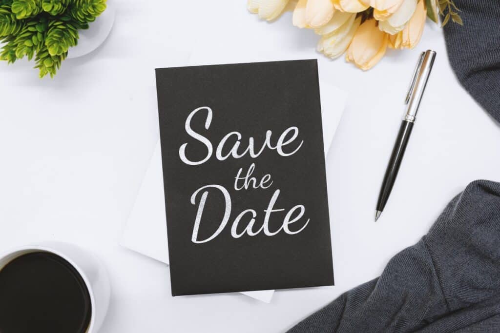 Save the date written on a black piece of paper on top of white table with pen next to it. 