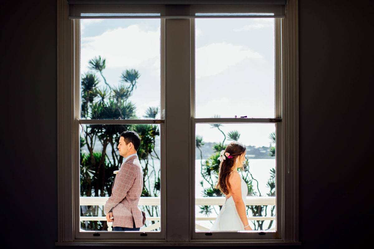 looking through a window and outside is the wedding couple not facing each other and about to turn around to see each other for first time. wedding couple