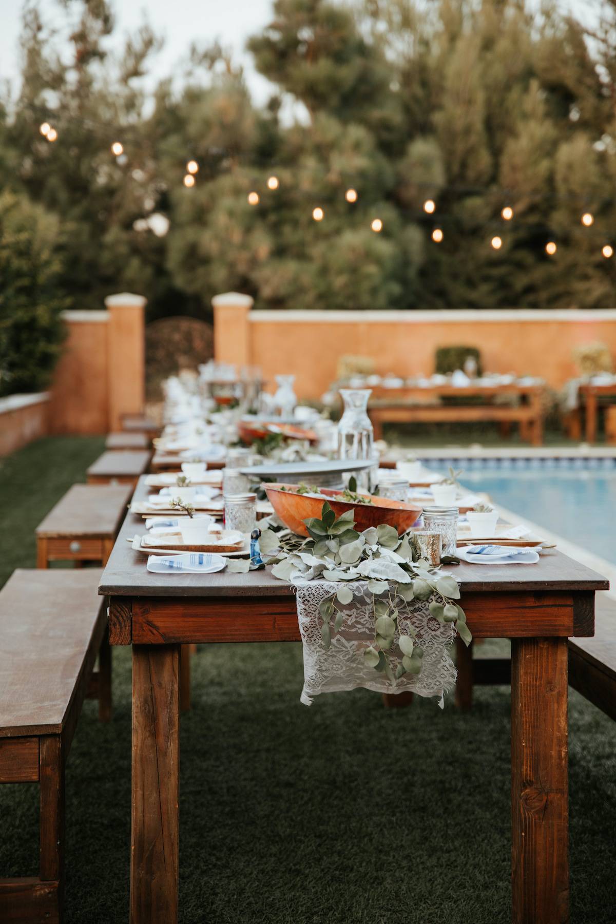 Backyard wedding setting - wooden tables with bohemian decor around swimming pool.