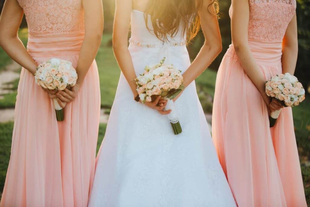The bride and bridesmaids in an elegant dress is standing and holding hand bouquets of pastel pink flowers and greens with ribbon at nature. Young beautiful girls holds a wedding bouquet outdoors..