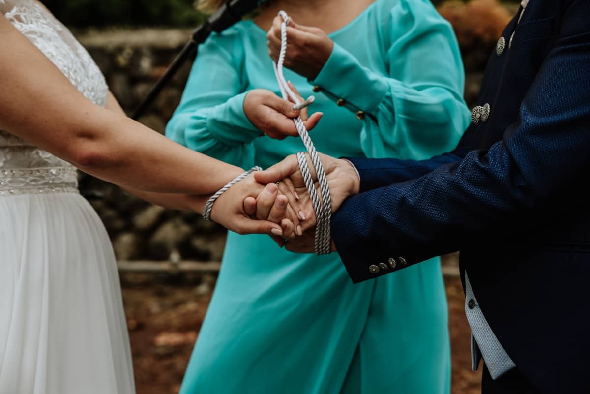 Bride and groom stand face to face with hands outstretched, a lady in a green dress stands between them binding their hands with grey handfasting cord